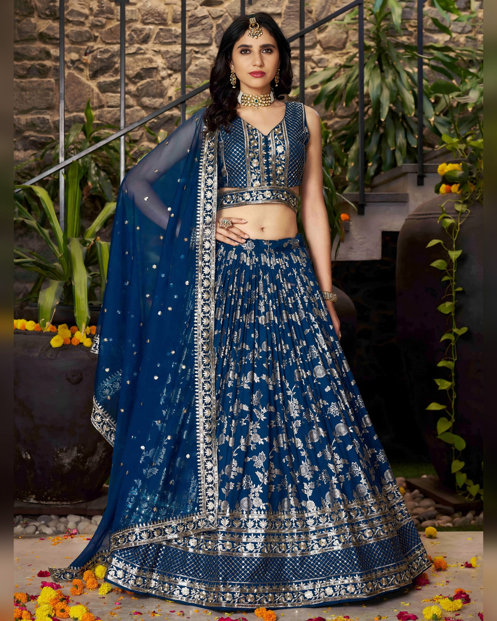 Woman in a blue embroidered traditional outfit with a matching dupatta against a stone wall and plant background.