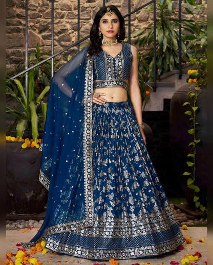 Woman in a blue embroidered traditional outfit with a matching dupatta against a stone wall and plant background.