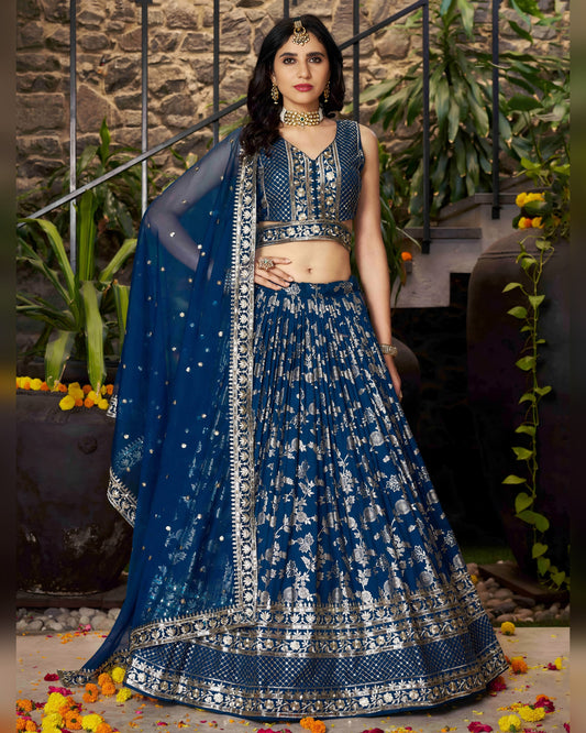 Woman in a blue embroidered traditional outfit with a matching dupatta against a stone wall and plant background.