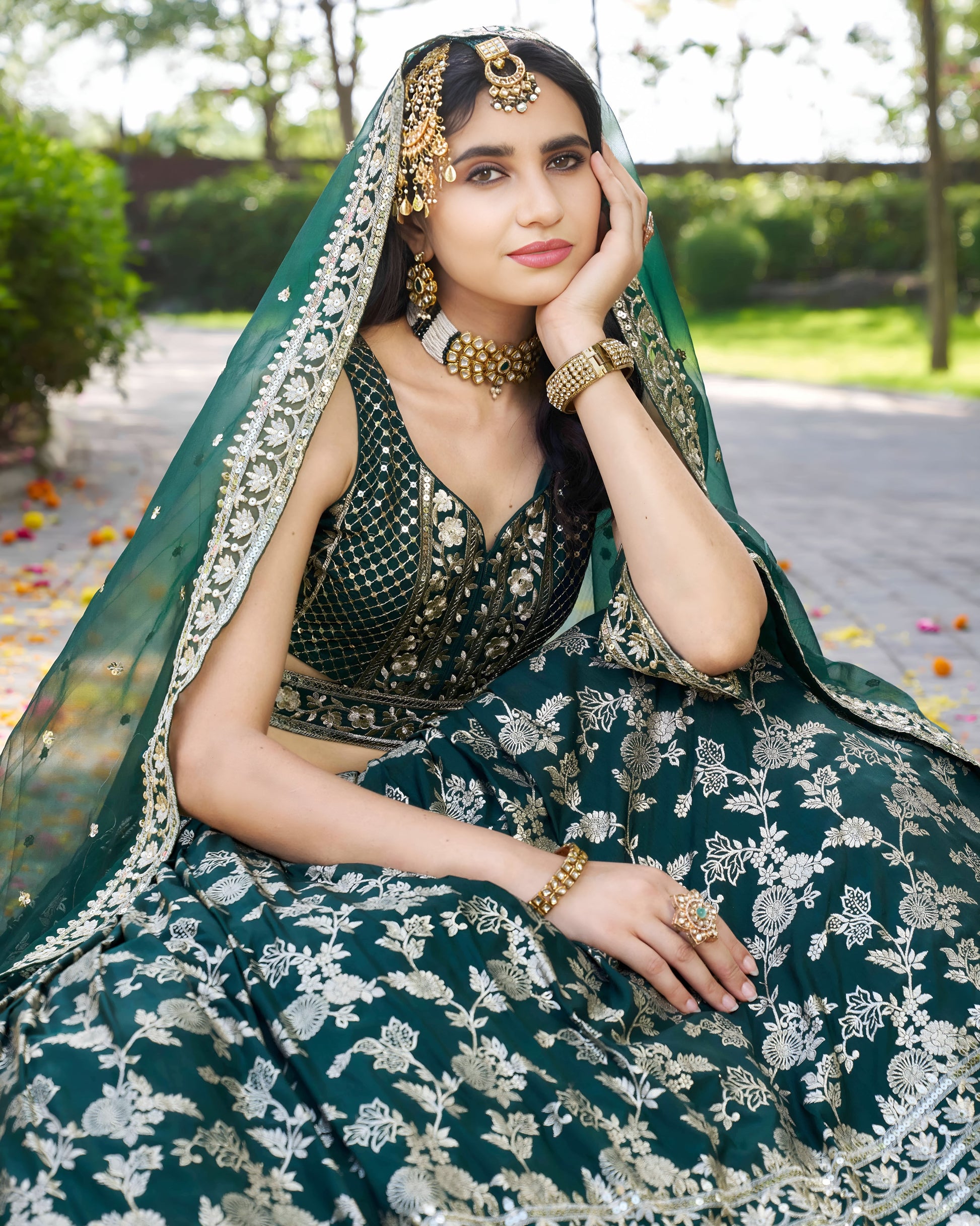 Woman in traditional green and gold outfit with jewelry, sitting outdoors.