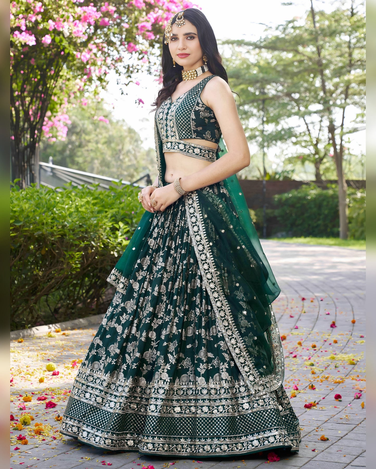 Woman in a green and black embroidered traditional outfit standing outdoors with flowers on the ground.