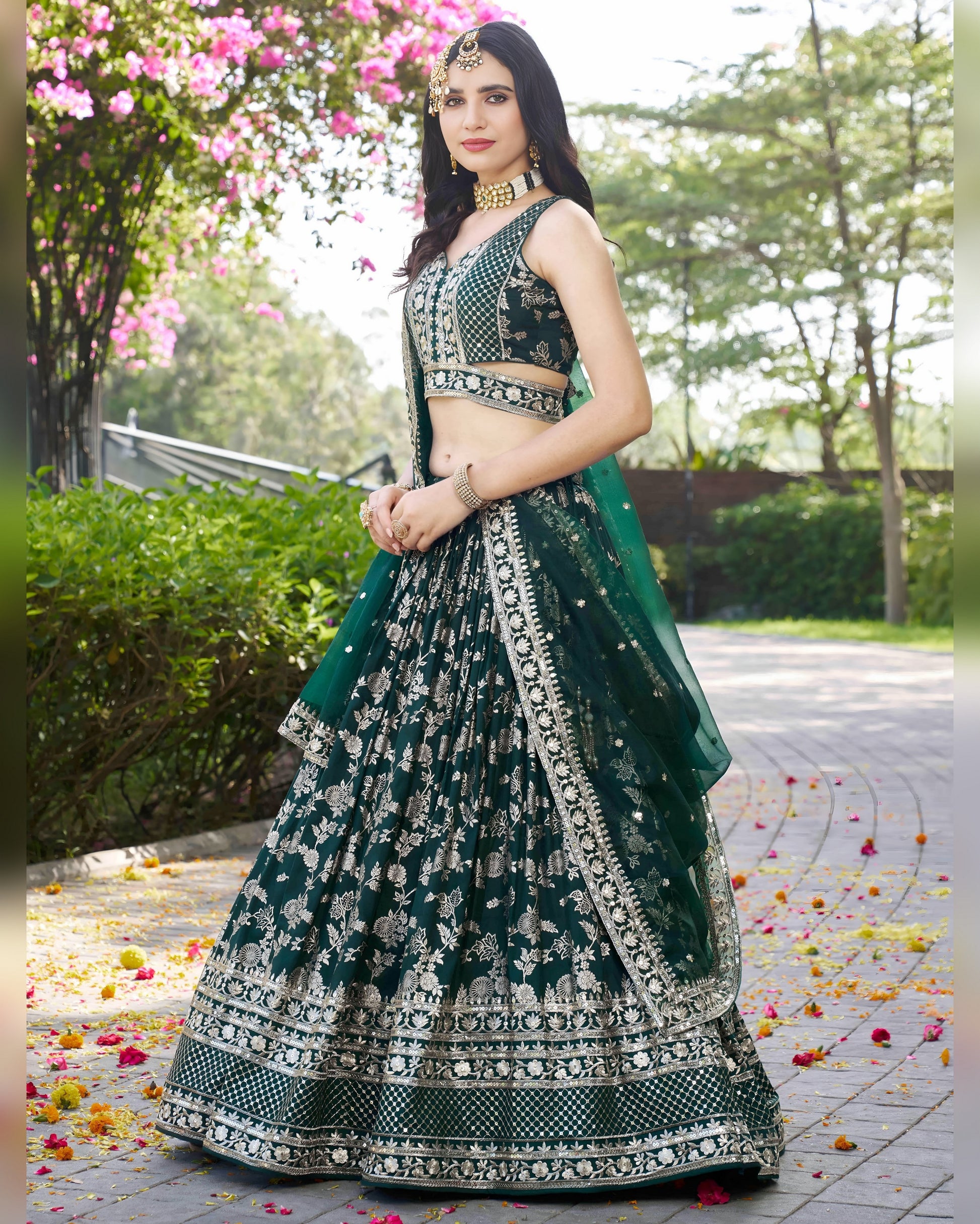 Woman in a green and black embroidered traditional outfit standing outdoors with flowers on the ground.