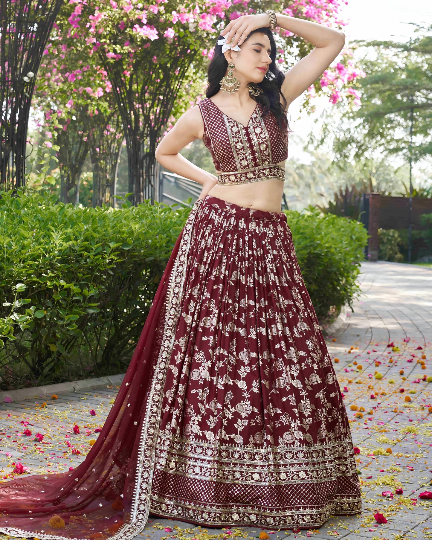 Woman in a maroon embroidered traditional outfit standing outdoors with greenery and flowers.