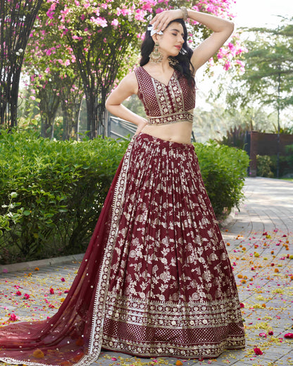 Woman in a maroon embroidered traditional outfit standing outdoors with greenery and flowers.