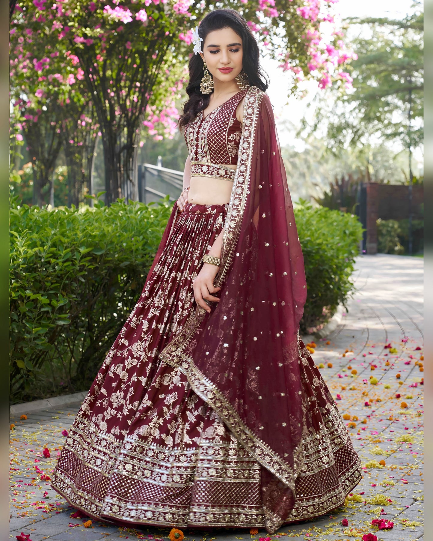 Woman in a maroon and gold traditional outfit standing outdoors with greenery and flowers in the background.