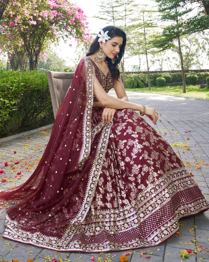 Woman in a maroon and gold traditional outfit sitting outdoors with flowers and greenery.