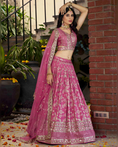 Woman in a pink traditional outfit standing against a brick wall with plants.