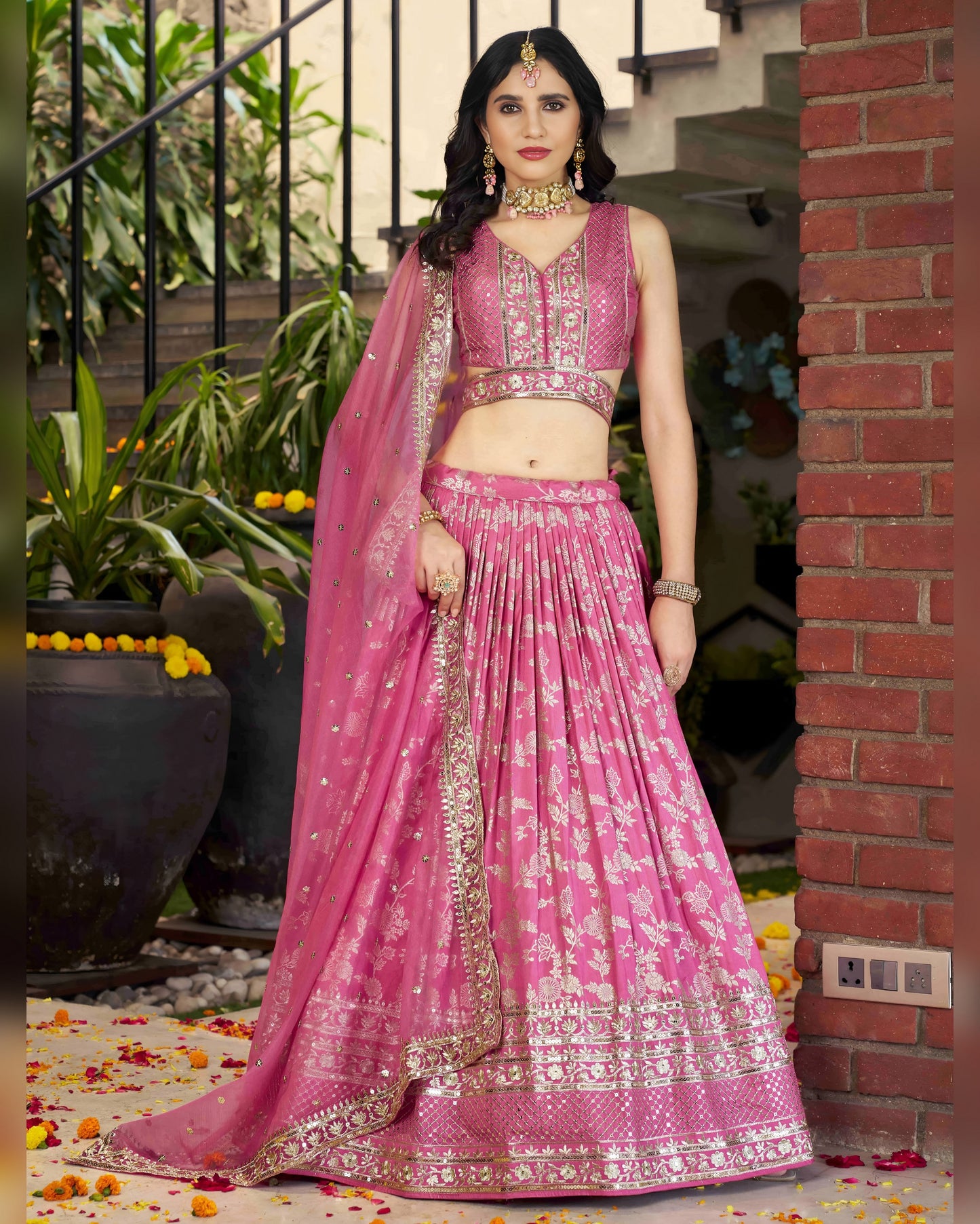 Woman in a pink traditional outfit standing outdoors with plants and a brick wall in the background.