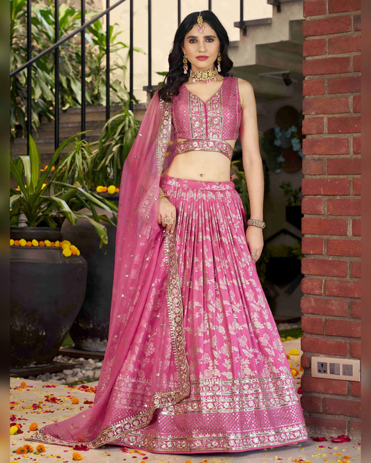 Woman in a pink traditional outfit standing outdoors with plants and a brick wall in the background.