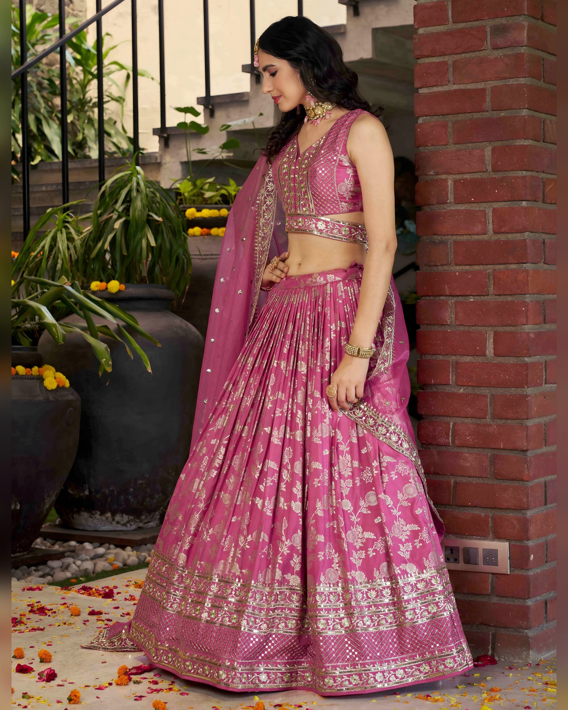 Woman in a pink traditional outfit standing against a brick wall with plants in the background.