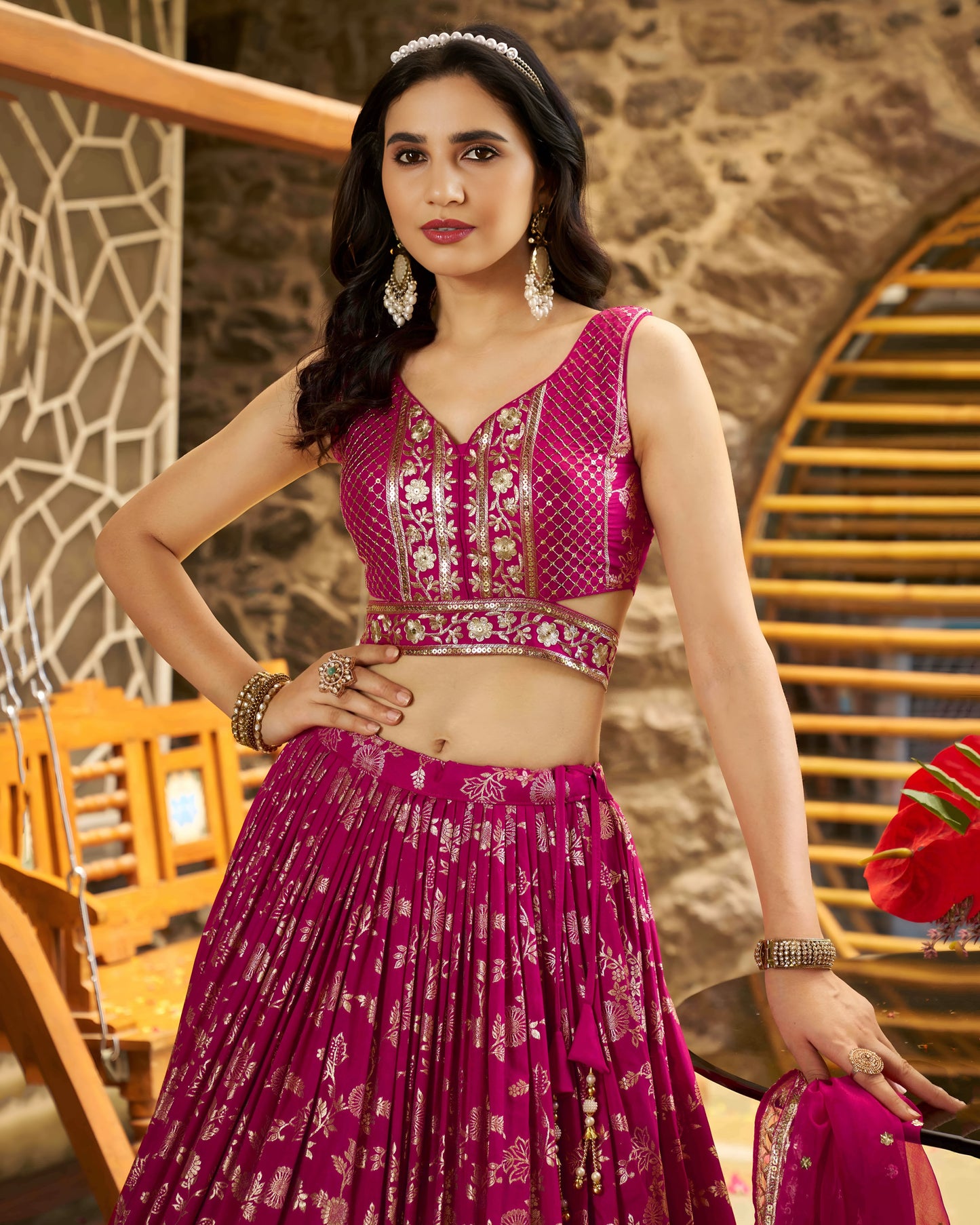 Woman in a pink traditional outfit with intricate patterns, standing against a stone wall.