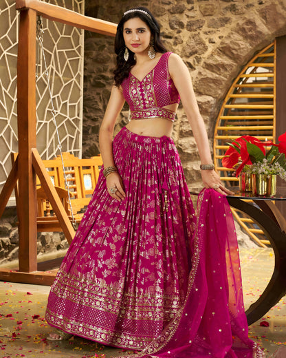 Woman in a pink traditional outfit standing in a decorative setting with flowers and furniture.