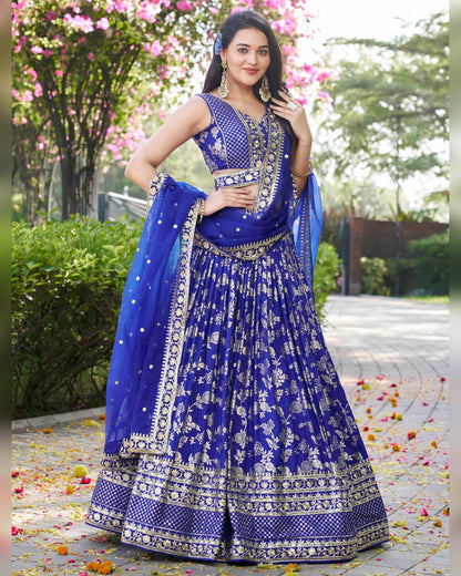 Woman in a blue embroidered traditional outfit standing outdoors with greenery and flowers in the background