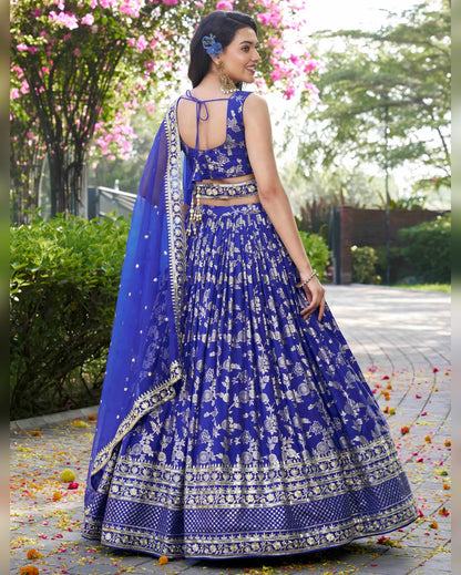 Woman in a blue and white embroidered traditional outfit standing outdoors with greenery in the background.
