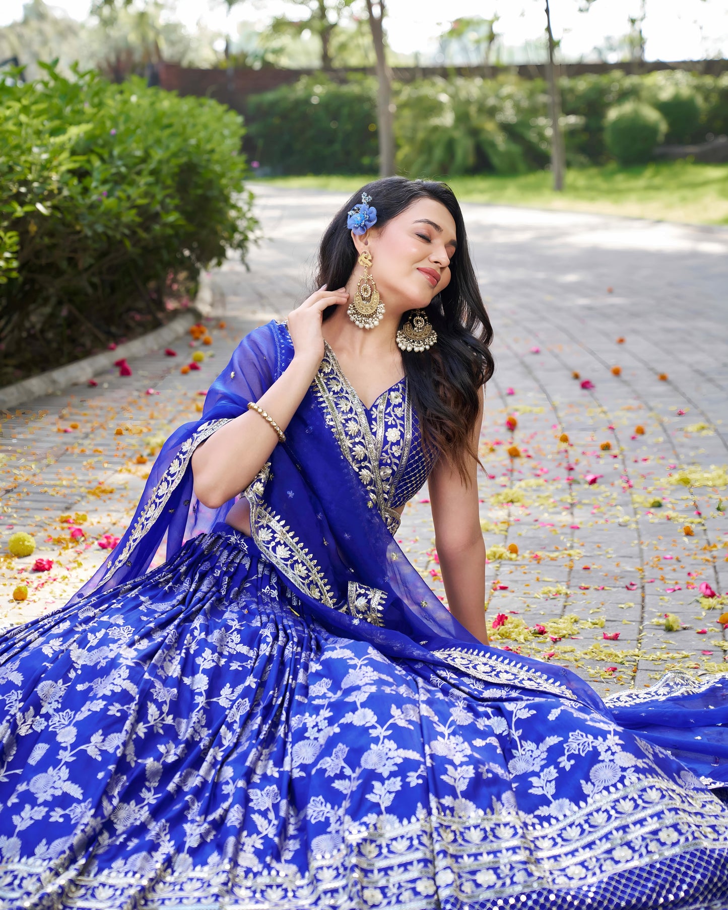 Woman in a blue and white traditional outfit sitting outdoors with greenery in the background