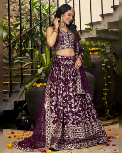 Woman in a purple traditional outfit with floral patterns, standing in an indoor setting with plants and stairs.