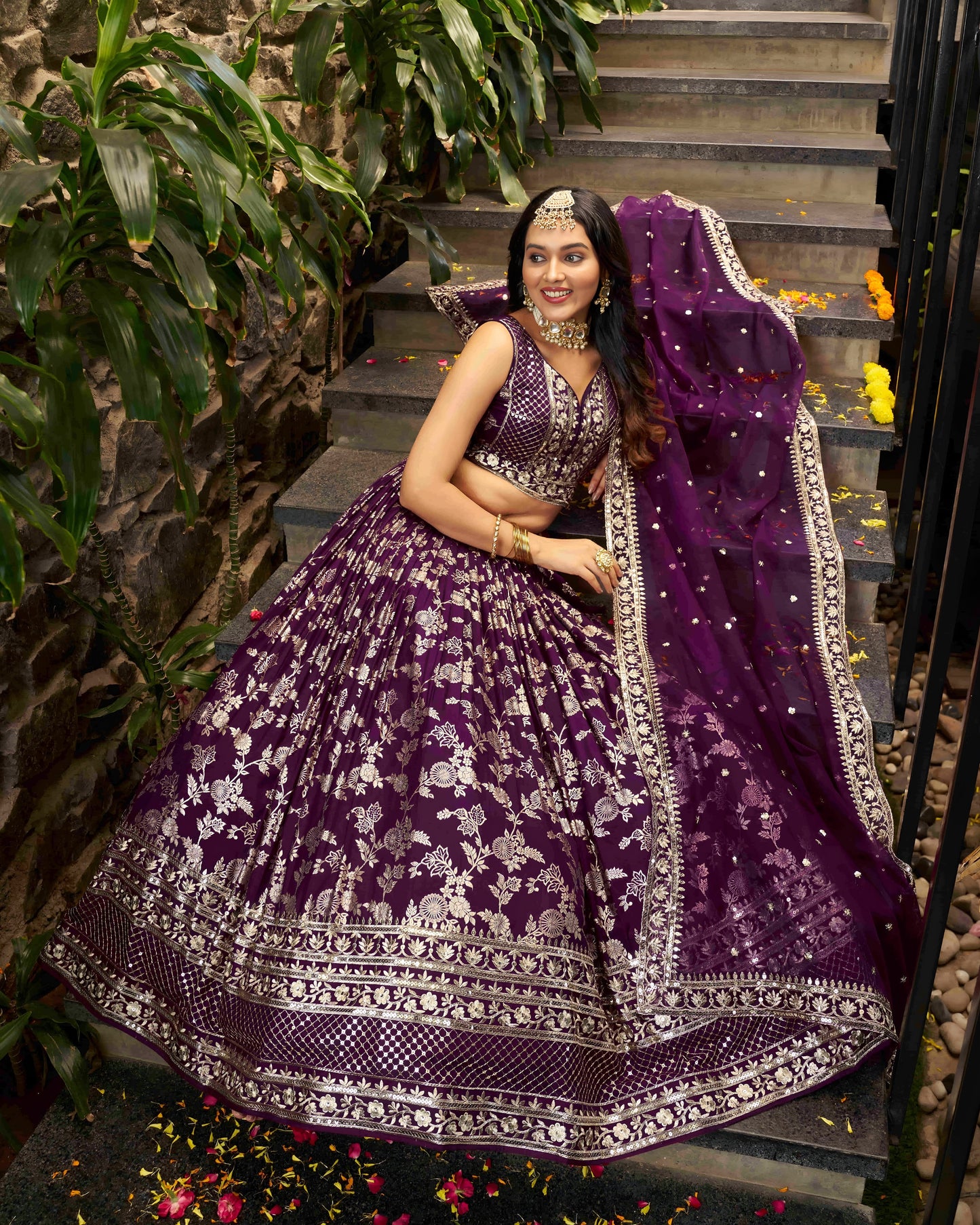 Woman in a purple embroidered traditional outfit sitting on steps with greenery around.