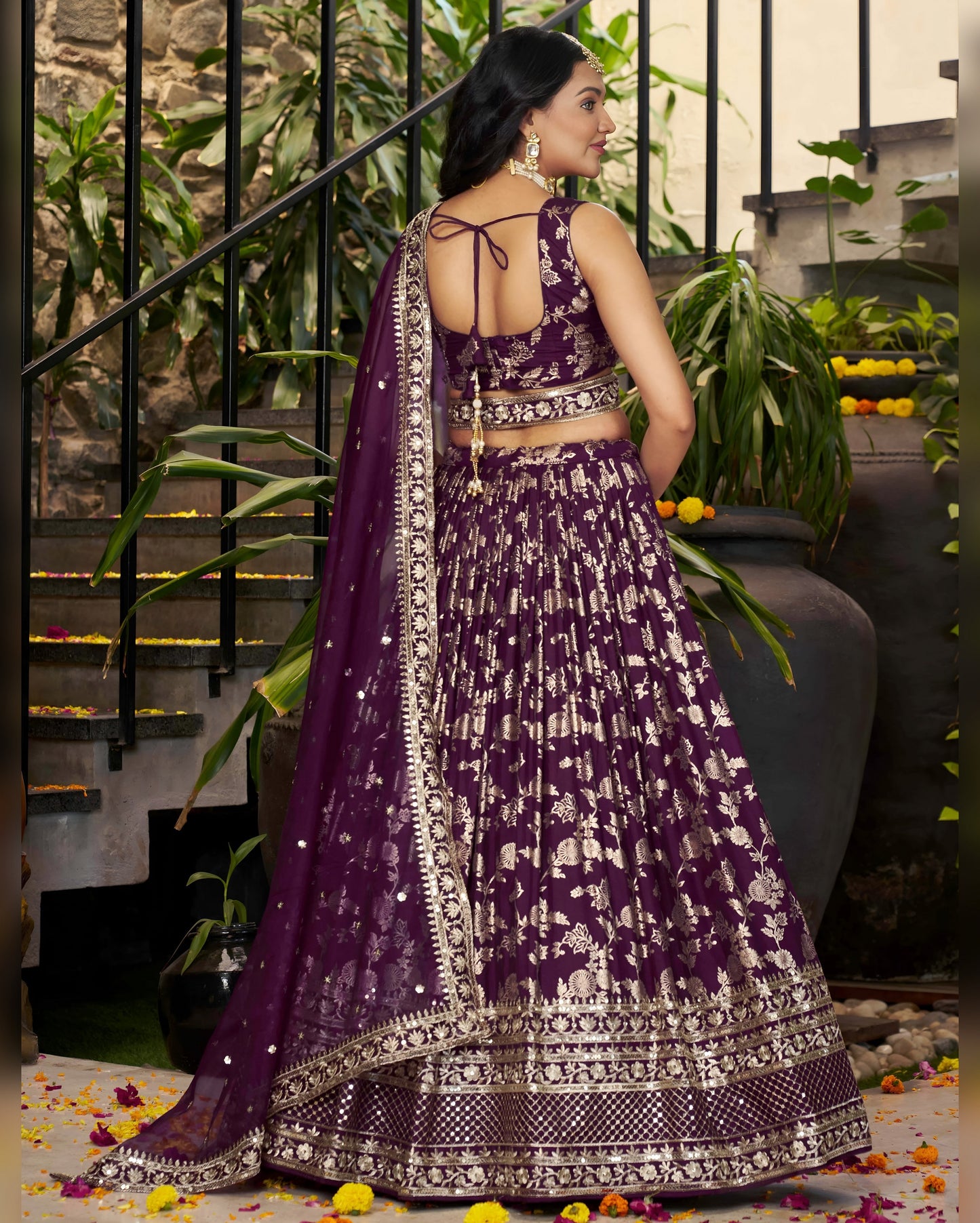 Woman in a purple and white traditional outfit standing in an outdoor setting with plants and stairs.