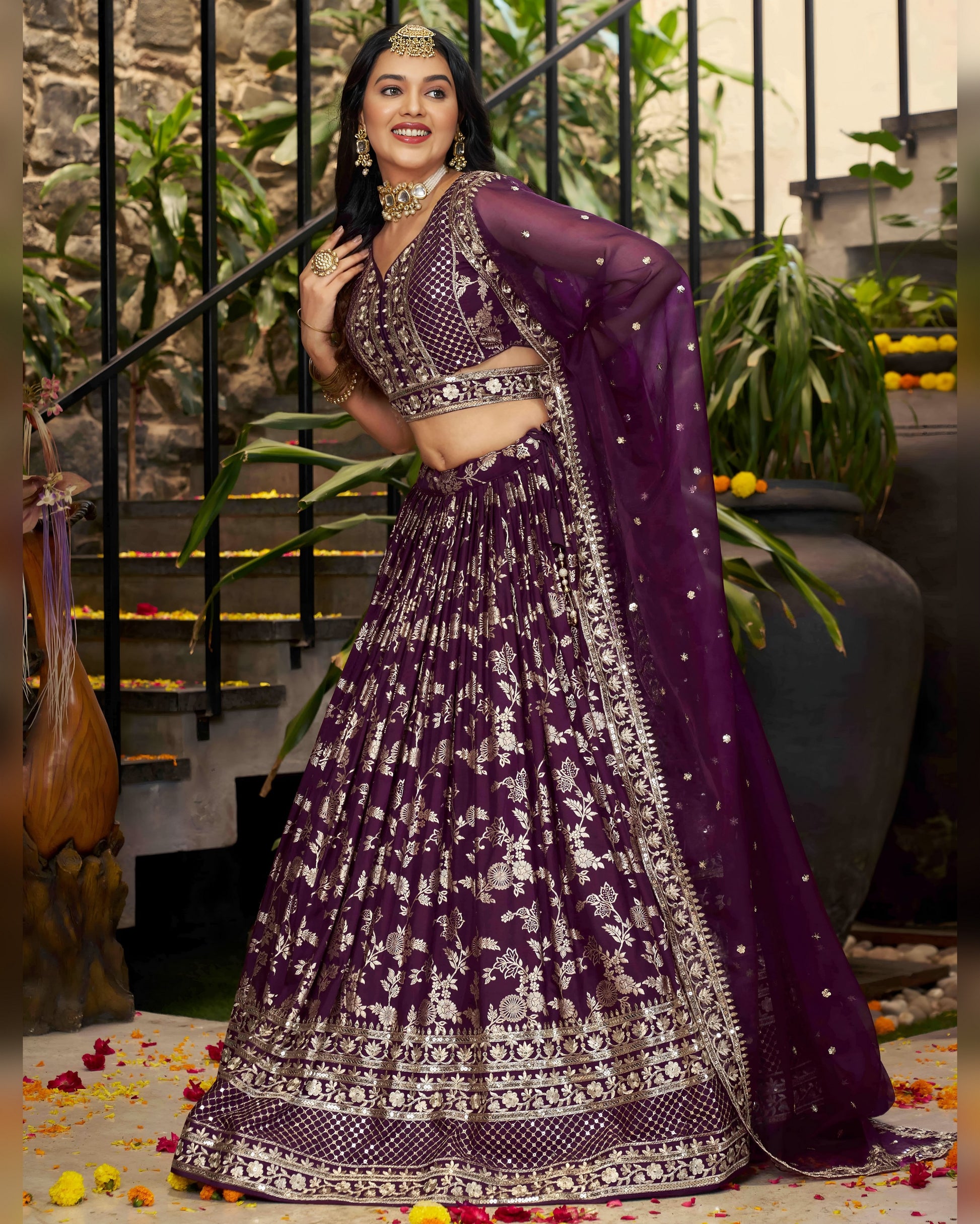 Woman in a purple traditional outfit with intricate designs, standing indoors with plants and decor.
