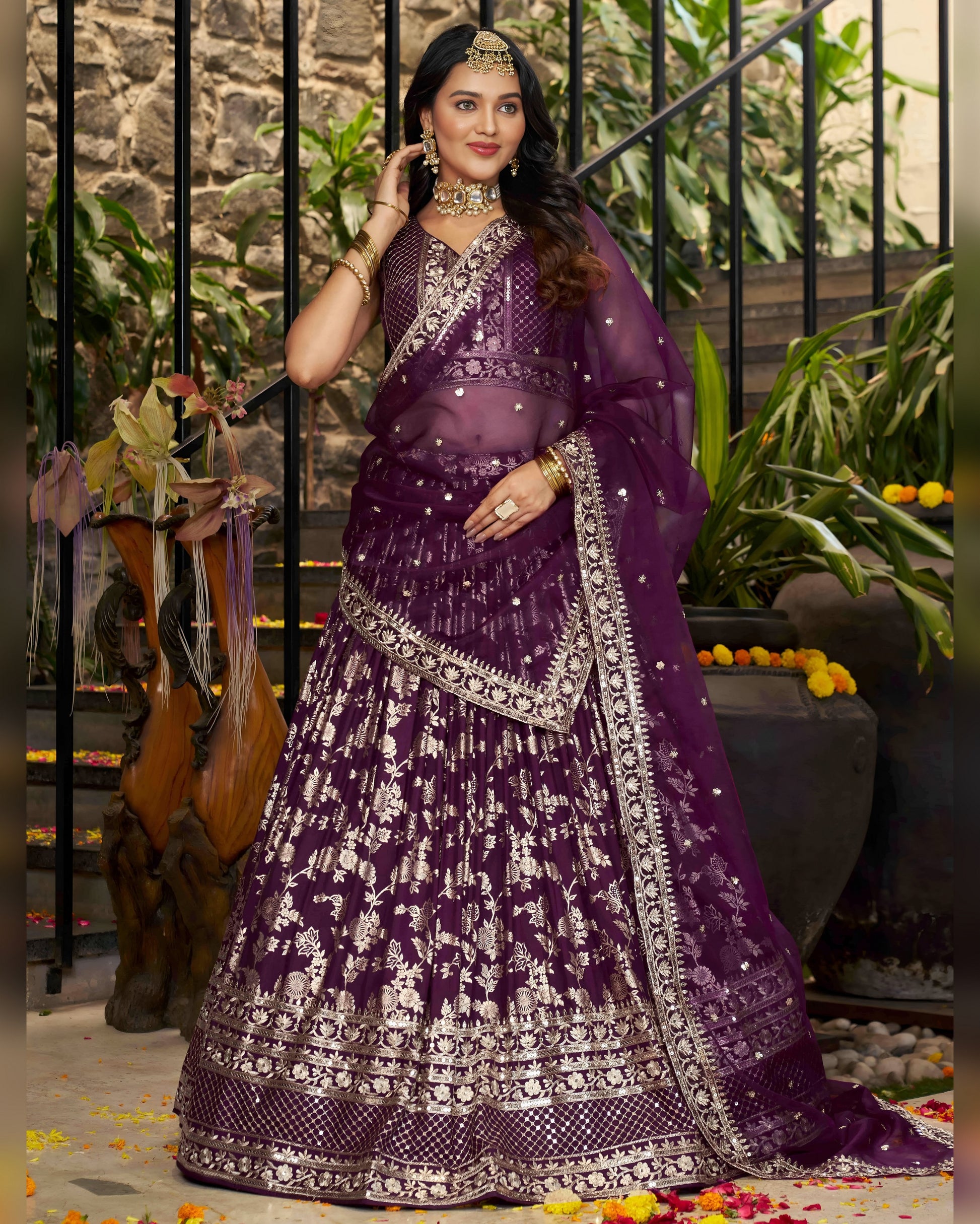 Woman in a purple traditional outfit with intricate designs, standing in an outdoor setting.