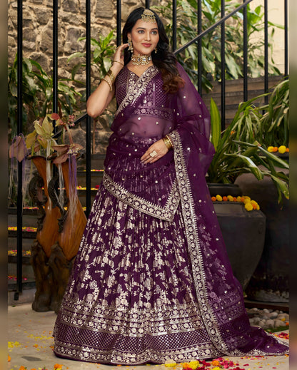 Woman in a purple traditional outfit with intricate designs, standing in an outdoor setting.