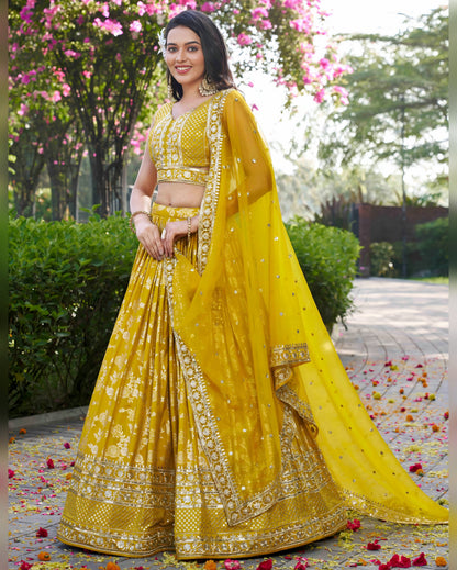 Woman in a yellow traditional outfit standing outdoors with greenery and flowers in the background