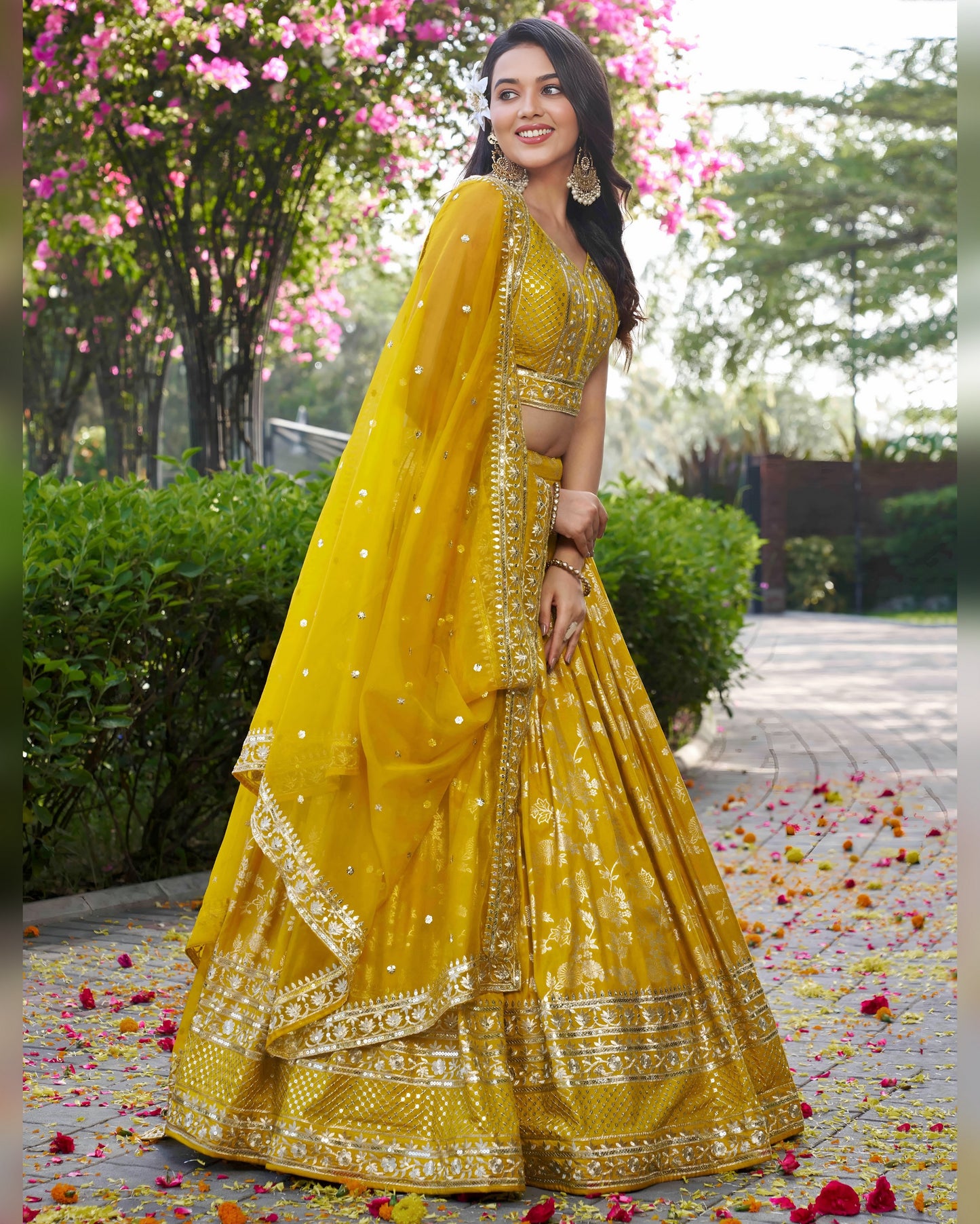 Woman in a yellow traditional outfit standing outdoors with flowers and greenery.