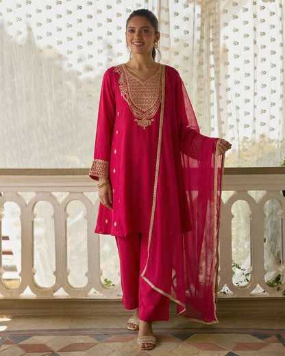Woman in a pink traditional outfit with gold embroidery standing in front of a decorative white curtain.
