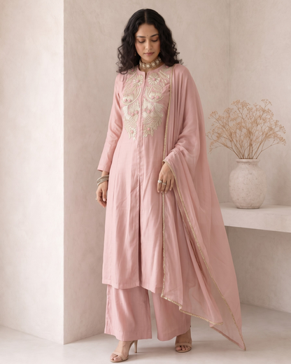 A woman models a light pink traditional Indian outfit consisting of a long kurta with gold embroidery, matching trousers, and a sheer dupatta, set against a minimalist beige wall with a decorative vase.