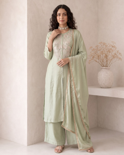 A woman models a light sage green traditional Indian salwar kameez suit with matching dupatta. The kurta features intricate gold embroidery on the chest, and the set is styled with a pearl necklace and neutral sandals in a minimalist beige interior setting.