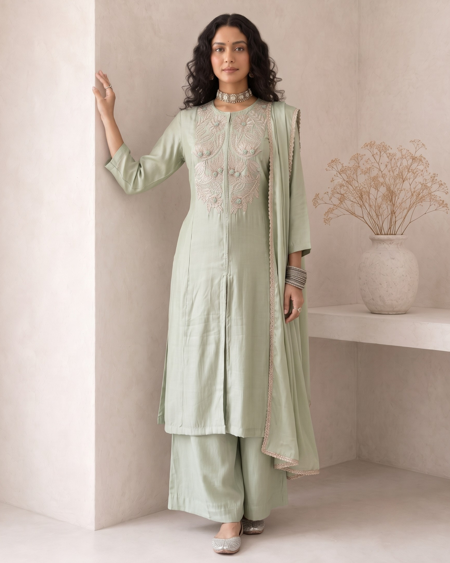 A woman models a light sage green three-piece salwar kameez set with intricate embroidery on the chest, matching wide-leg trousers, and a draped dupatta, set against a minimalist beige wall with a decorative vase.