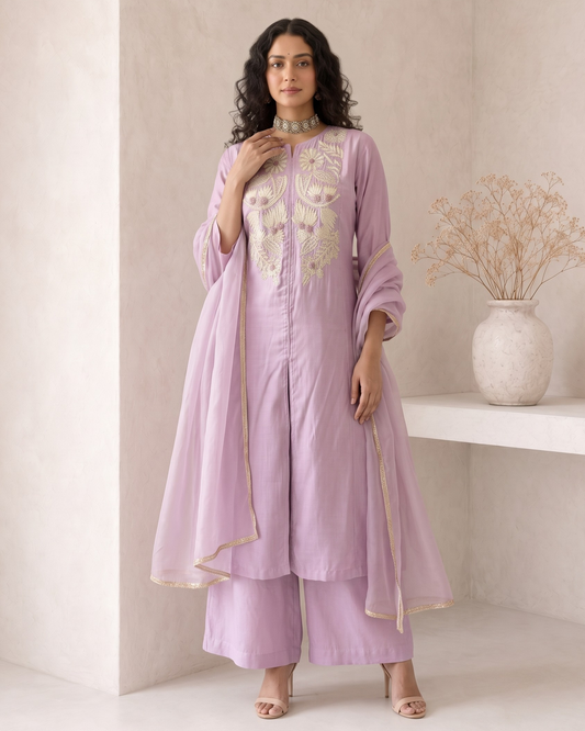 A woman models a lavender purple traditional Indian outfit consisting of a kurta with cream embroidery, matching wide-leg pants, and a sheer dupatta, set against a minimalist beige wall with a decorative vase.