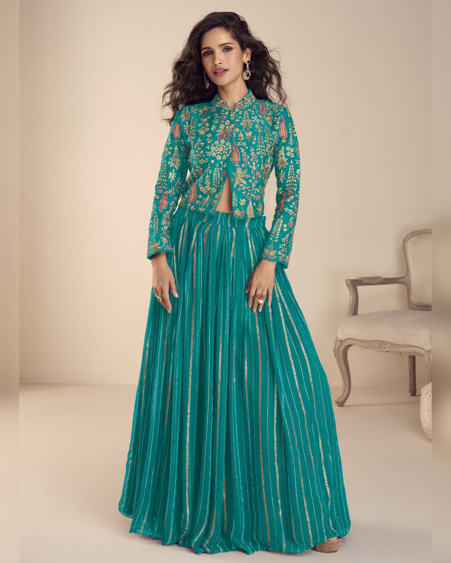 Woman wearing a teal embroidered outfit in a minimalistic room.