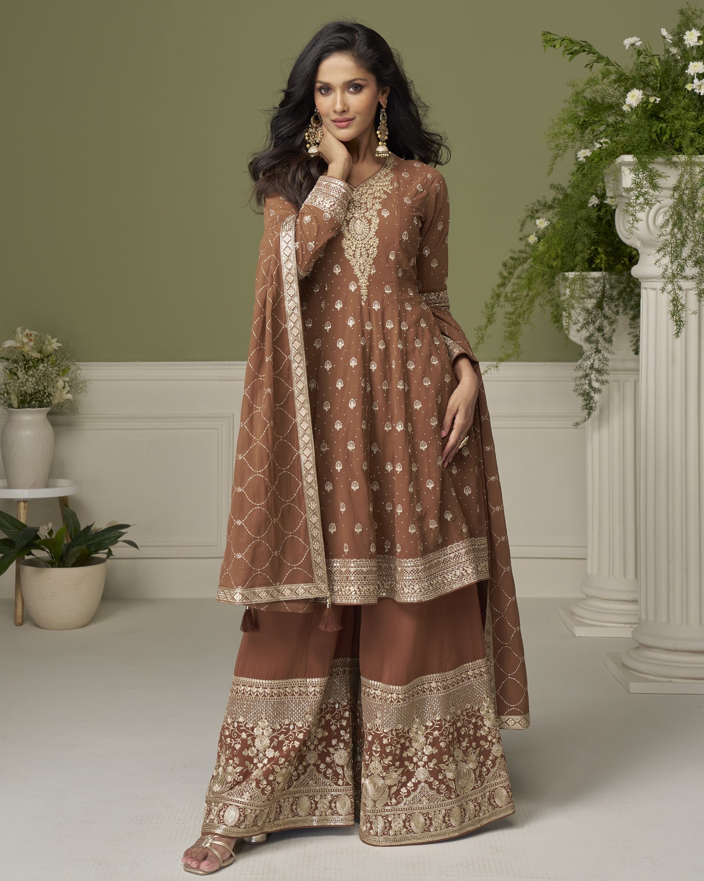 Woman in a brown embroidered traditional outfit standing in a decorated room with plants.