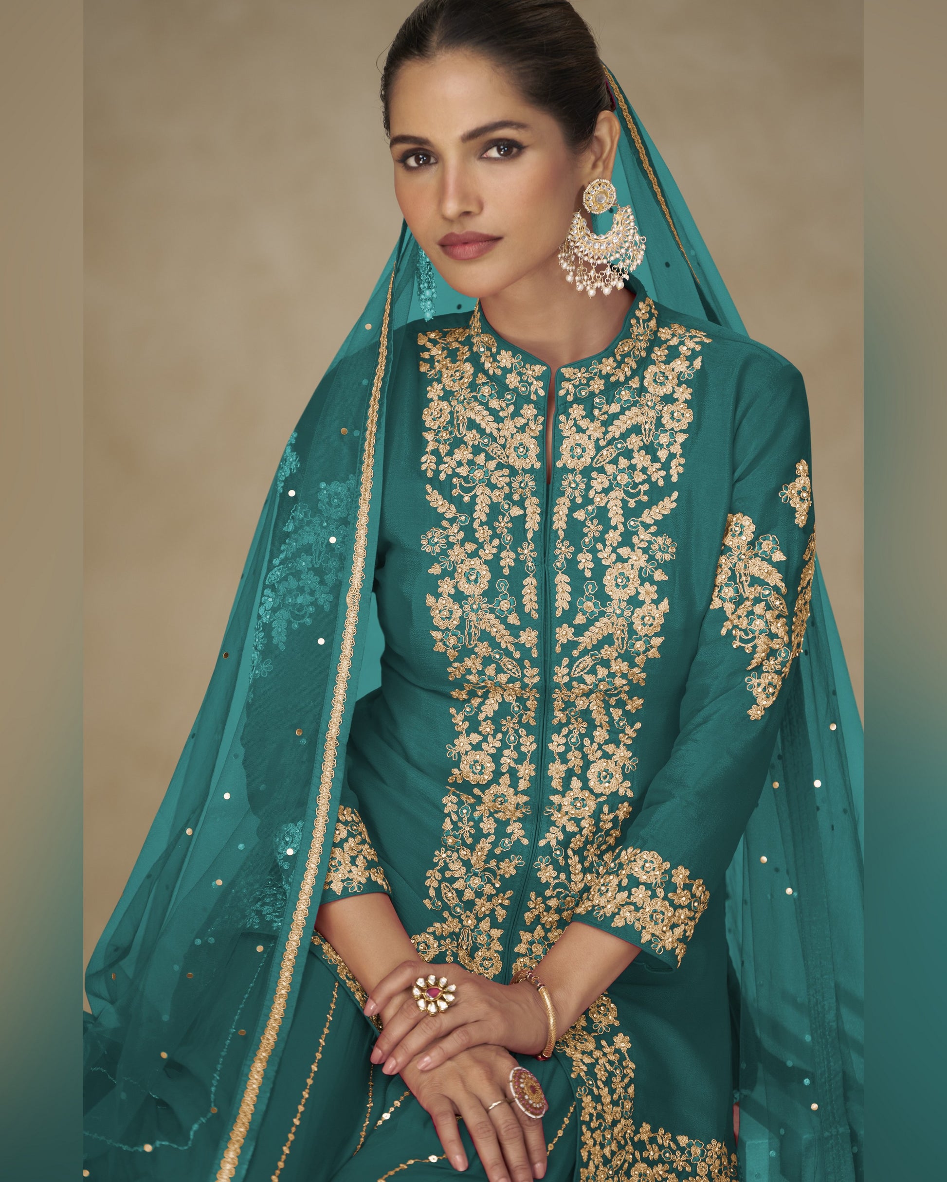 Woman wearing a teal traditional outfit with gold embroidery against a beige background