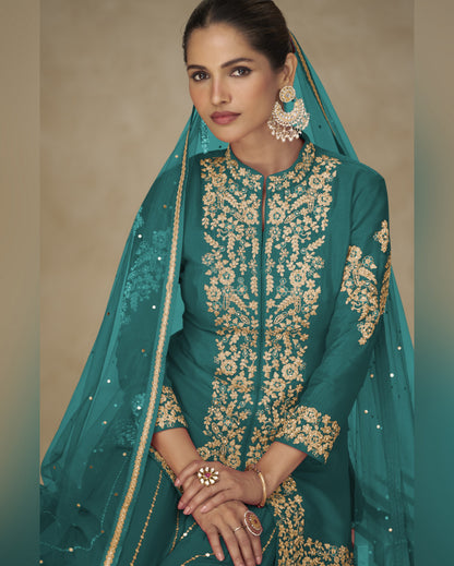 Woman wearing a teal traditional outfit with gold embroidery against a beige background