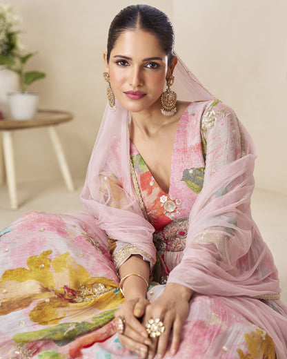 Woman in a floral dress with a pink dupatta sitting indoors.