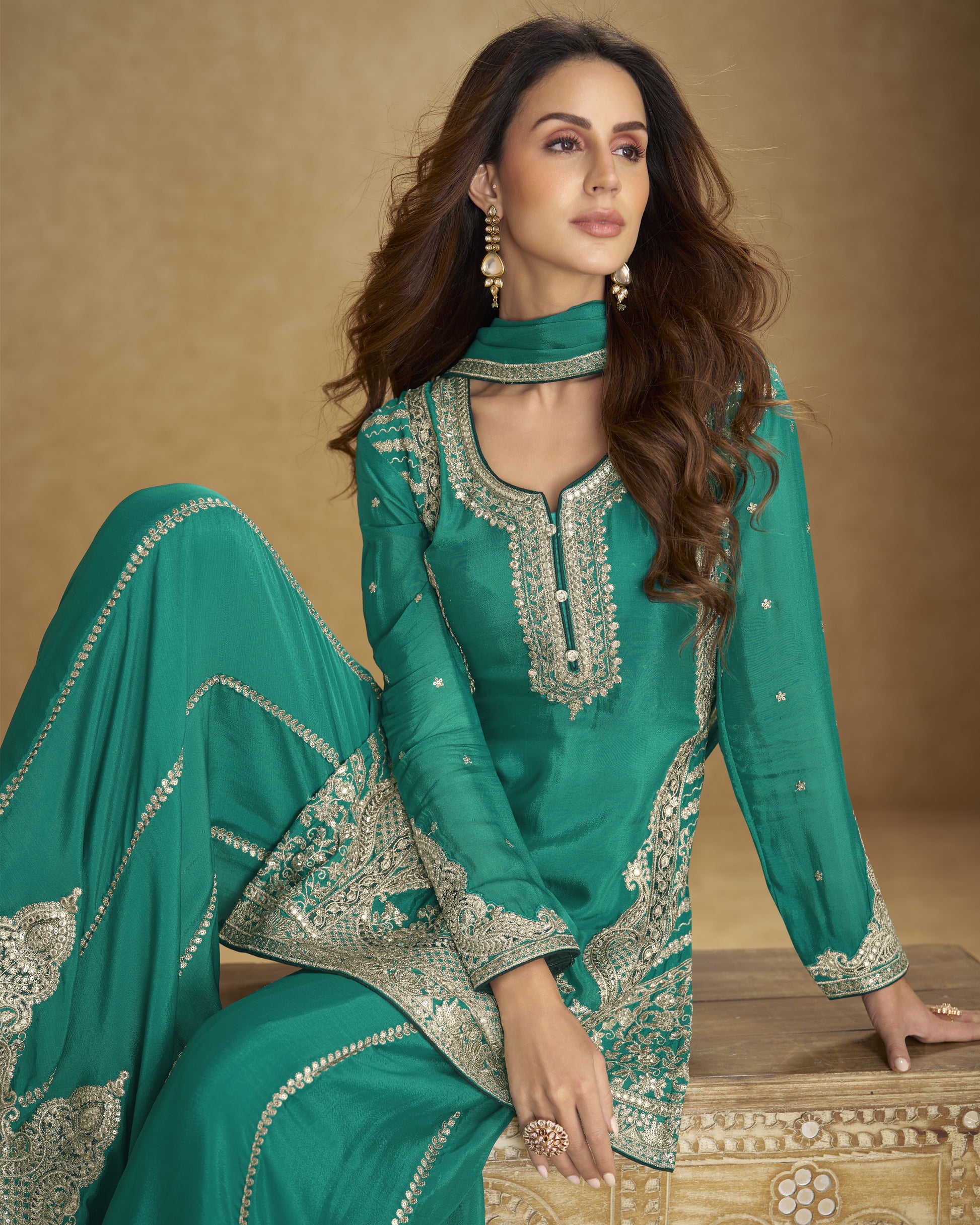 Woman in a teal embroidered traditional outfit sitting on a wooden stool against a beige background
