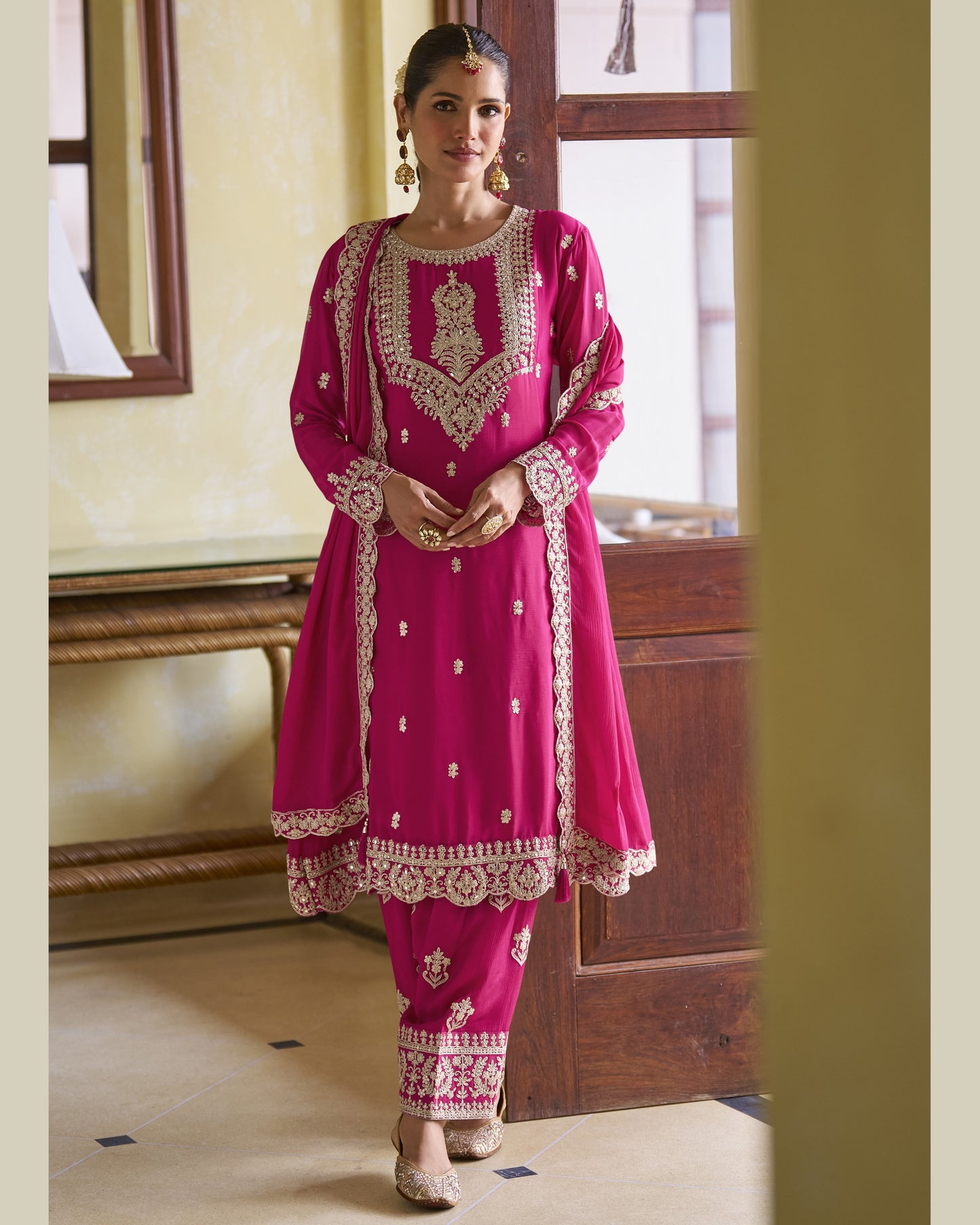 Woman wearing a pink traditional outfit with intricate embroidery in a room.