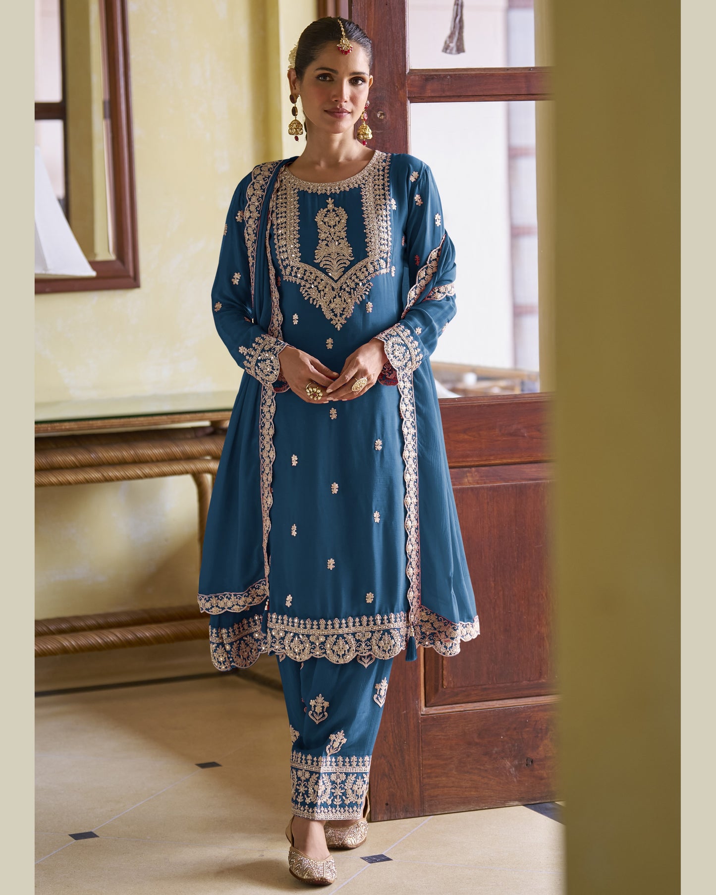 Woman wearing a blue embroidered traditional outfit in a room with wooden furniture.