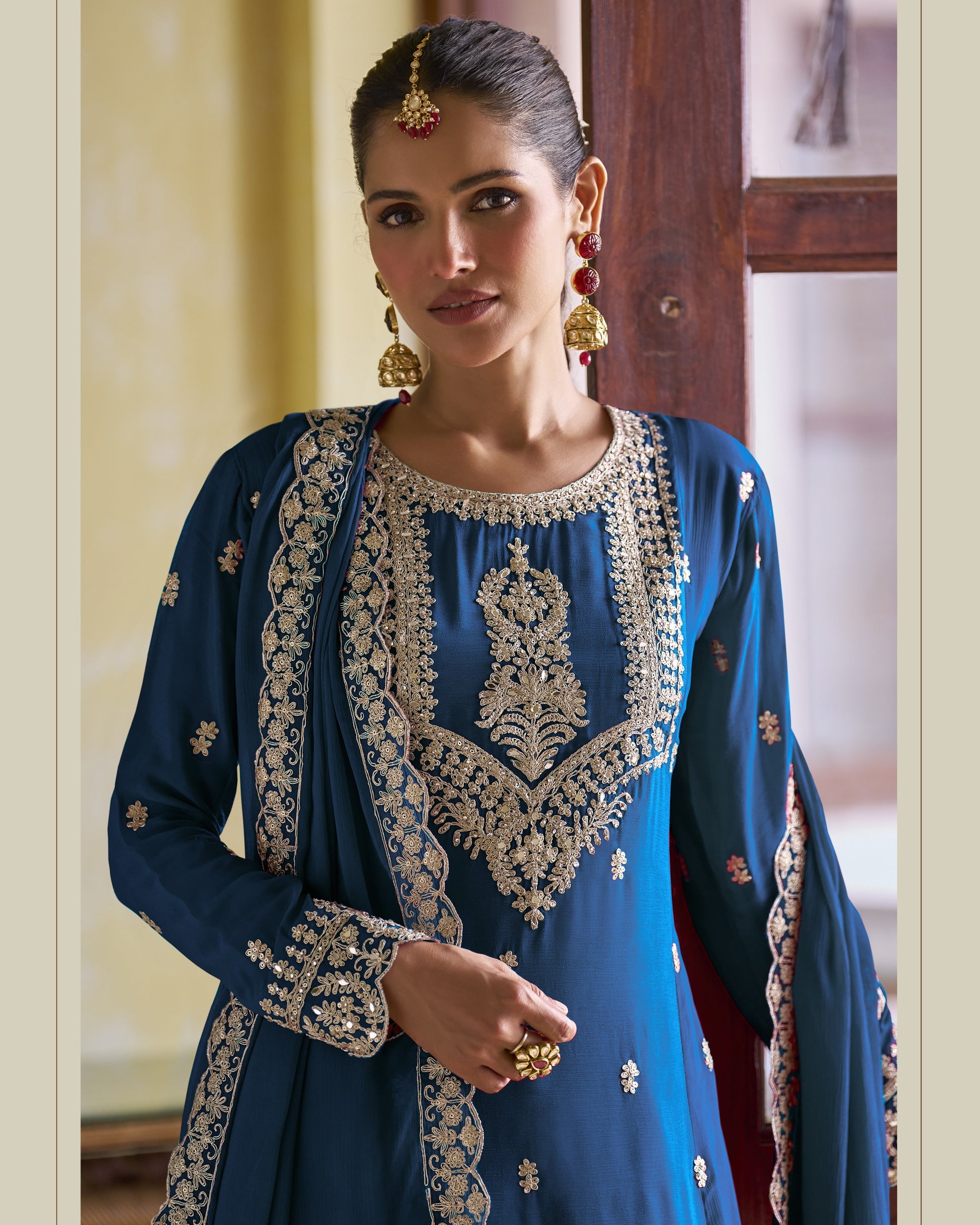 Woman wearing a blue embroidered traditional outfit with gold jewelry indoors.