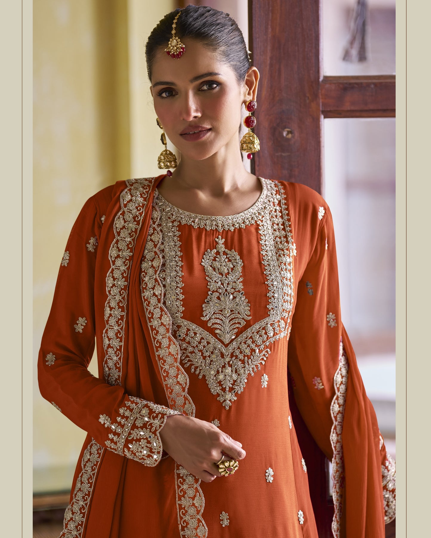 Woman wearing an orange traditional outfit with intricate white embroidery in a room setting.