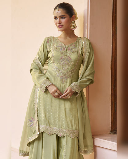 Woman wearing a light green embroidered traditional outfit against a beige wall.