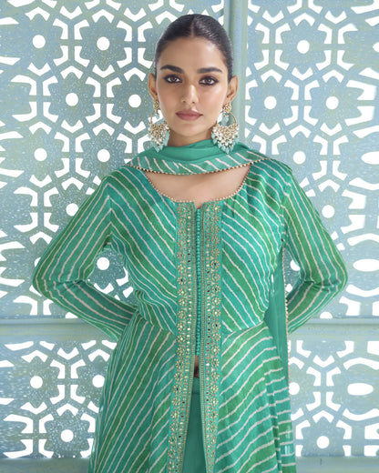 A woman modeling a green traditional outfit with vertical white stripes and gold embroidery, standing in front of a light blue geometric patterned screen.