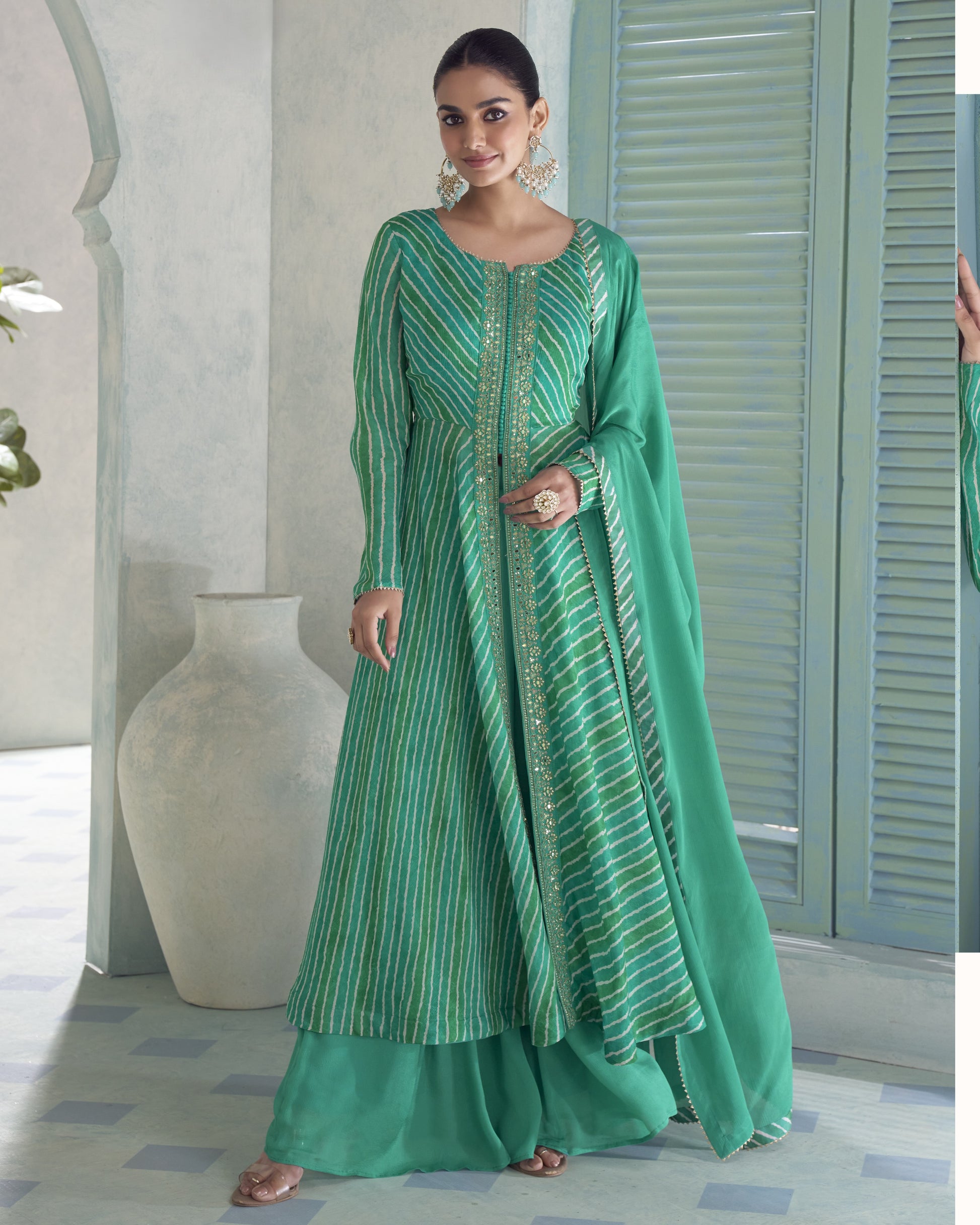 A woman models a green traditional Indian outfit with matching pants and a draped scarf, standing in a styled interior setting with a large vase and shuttered door in the background.