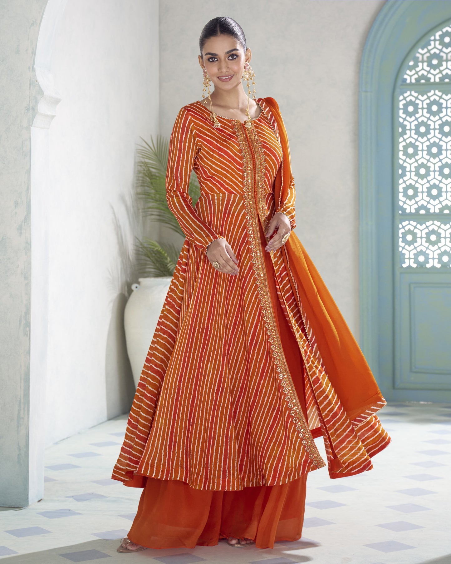 A woman modeling a traditional Indian outfit consisting of an orange and yellow striped kurta, matching orange pants, and a solid orange dupatta, set against a light-colored architectural background with blue arches.