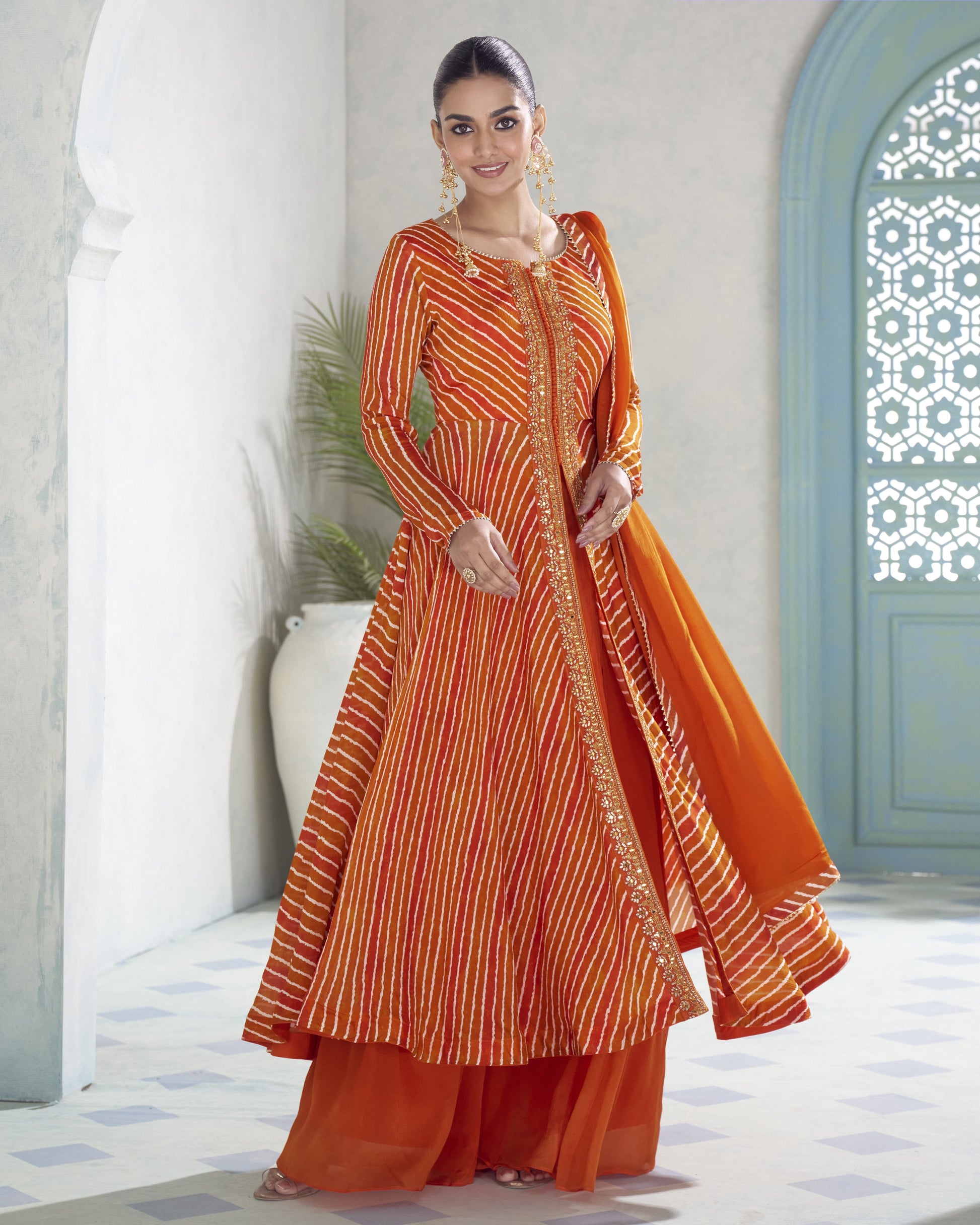 A woman modeling a traditional Indian outfit consisting of an orange and yellow striped kurta, matching orange pants, and a solid orange dupatta, set against a light-colored architectural background with blue arches.