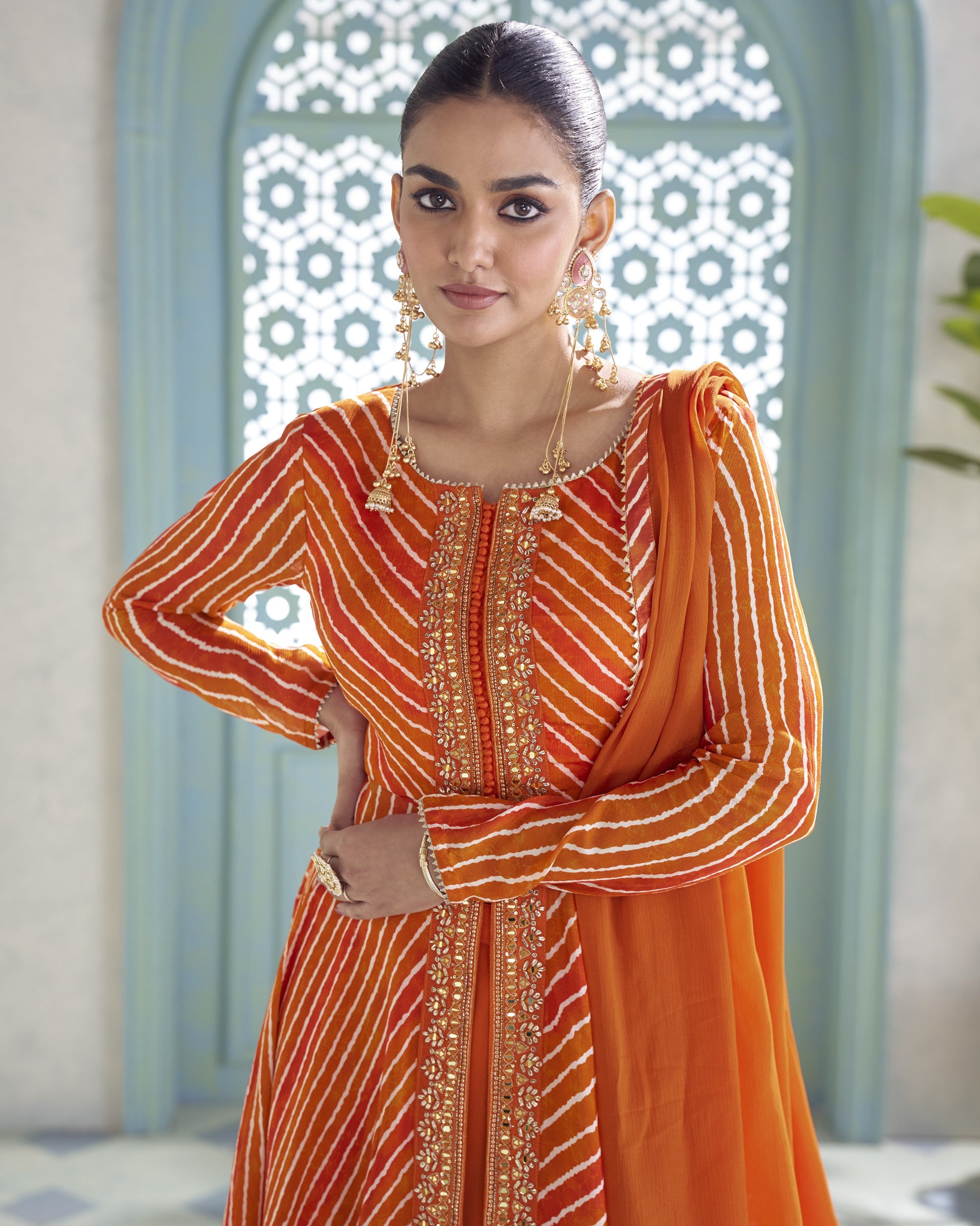 A woman modeling a traditional Indian outfit featuring a bright orange and red striped kurta with gold embroidery, paired with a matching orange dupatta, set against an ornate light blue archway.