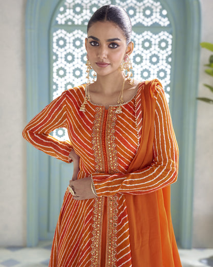 A woman modeling a traditional Indian outfit featuring a bright orange and red striped kurta with gold embroidery, paired with a matching orange dupatta, set against an ornate light blue archway.