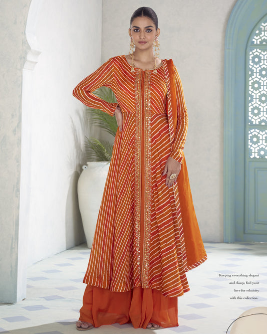 A woman models a traditional Indian outfit consisting of an orange and gold striped kurta with matching orange palazzo pants, standing in a brightly lit courtyard with white walls and a blue door.